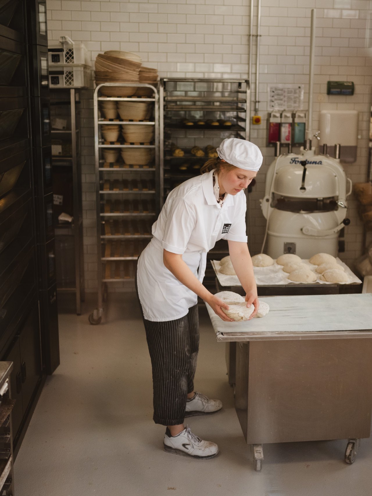 Eine Person in einem wei&szlig;en T-Shirt und einer Sch&uuml;rze legt Brot auf ein Backblech. Im Hintergrund stehen Regale mit Schalen und ein Knetger&auml;t.